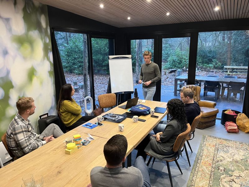 Een team van zes personen in gesprek rond de vergadertafel, met flip-over en uitzicht op het bos